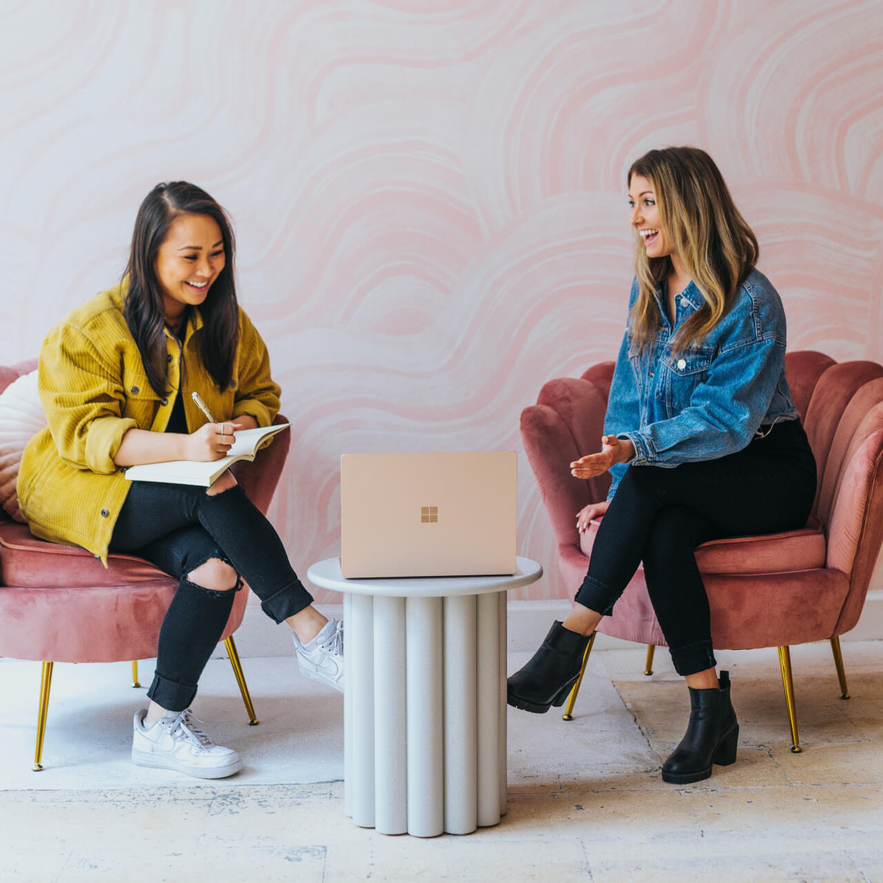 two young women are speaking and laughing while they are working on something and making notes