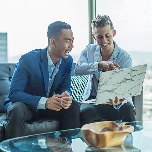 two men are sitting on the sofa and watching something on a laptop a glass table is in front of them